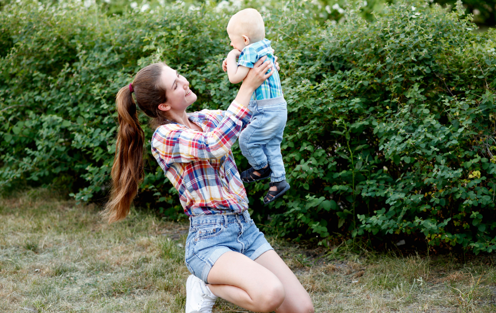 A woman kneels on grass and lifts a baby upward. Both are outdoors near green bushes, wearing plaid shirts and denim.