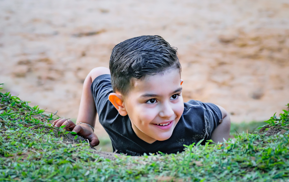 A young boy with short dark hair smiles while climbing up a grassy slope, with a sandy background visible behind him.