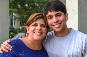 A woman and a young man stand close together, smiling at the camera. The woman has her arm around the young man's shoulder. They are outdoors near a building.