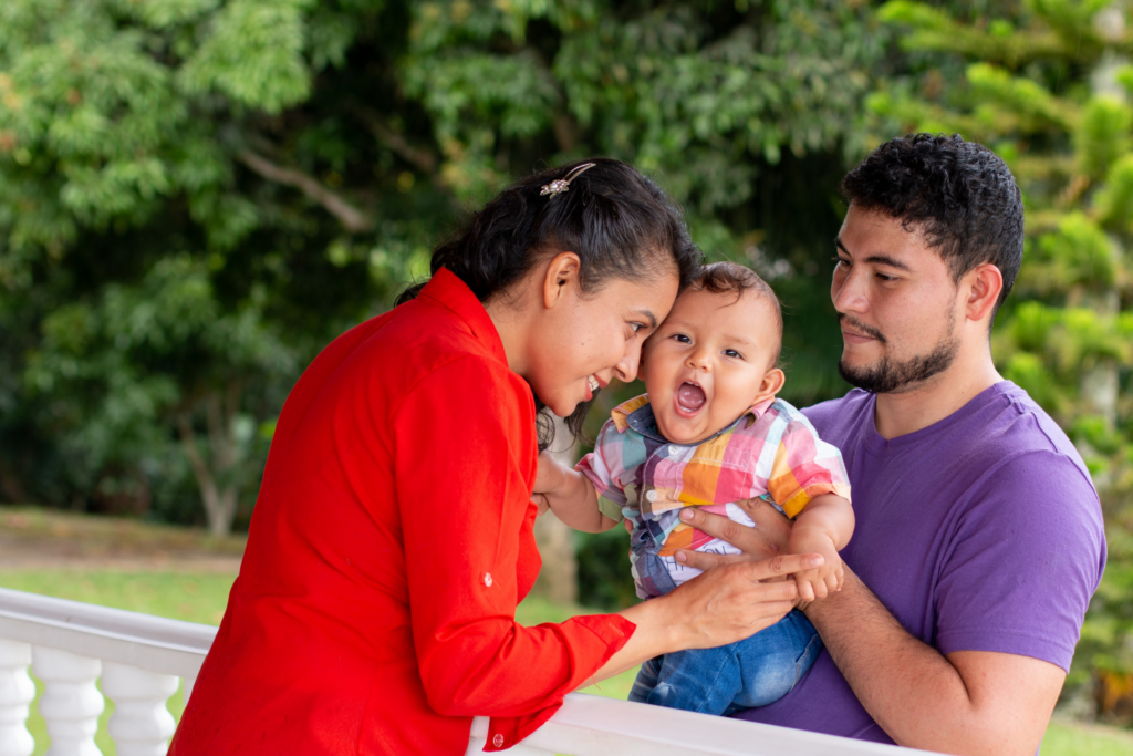 A woman in a red shirt interacts with a smiling baby held by a man in a purple shirt, standing outdoors near a white railing with trees in the background.