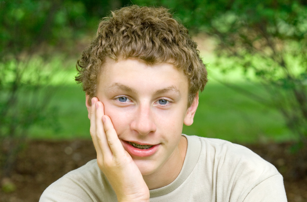 Teenage boy with light brown curly hair and braces resting his chin on his hand, sitting outdoors with green foliage in the background.