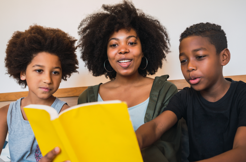 A woman sits between two children, holding a yellow book as they read together and look at the pages.