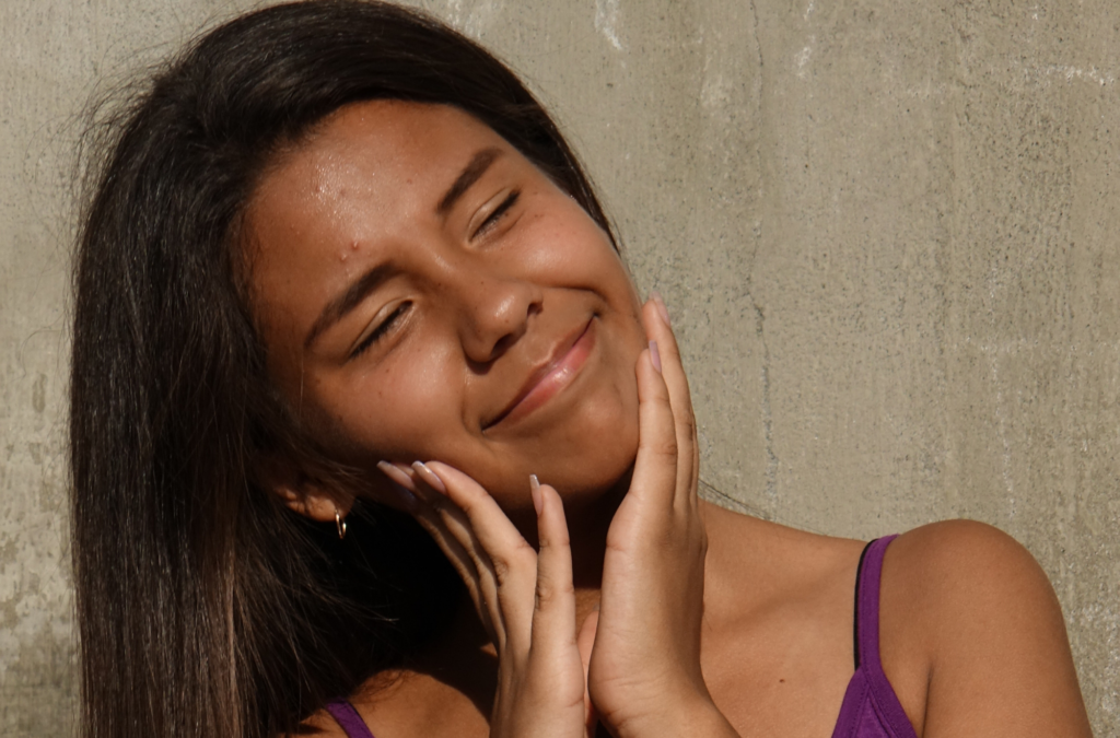 A young person with long dark hair smiles with eyes closed, gently touching their face with both hands, standing in sunlight against a plain concrete wall.
