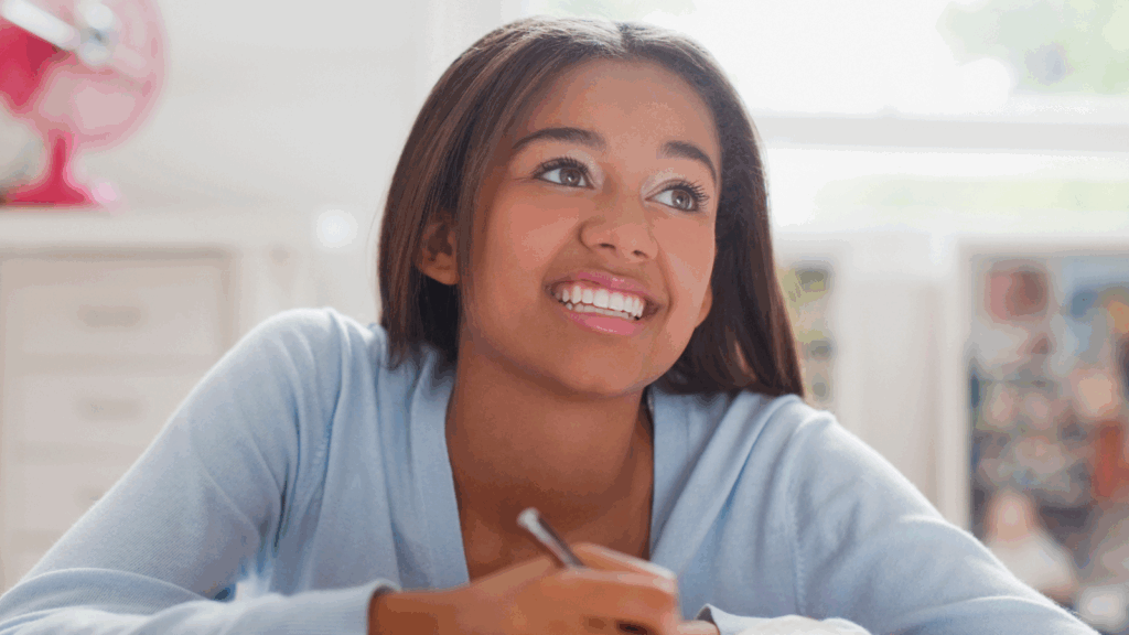 A young woman with long brown hair smiles as she writes with a pencil, sitting indoors in a bright room.