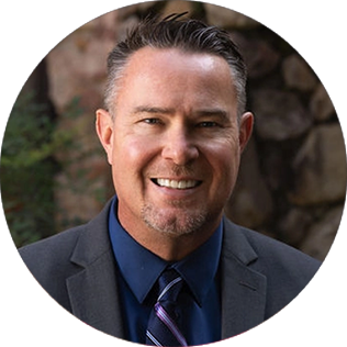 Man wearing a dark suit, blue shirt, and striped tie, smiling in an outdoor setting with a stone wall background.