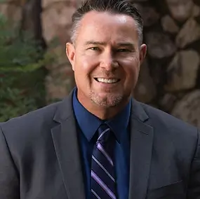 A man in a dark suit, blue shirt, and striped tie smiles at the camera with a stone wall and greenery in the background.