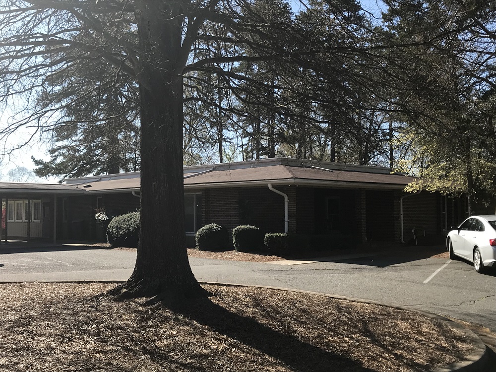 A single-story brick building with a brown roof, surrounded by trees and bushes. A white car is parked to the right on a paved driveway.