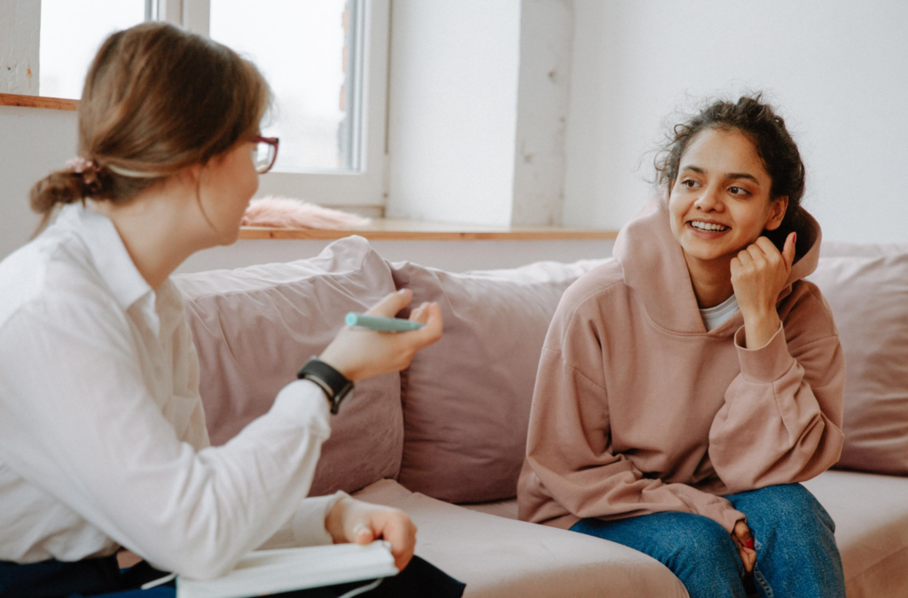Two women sit on a couch having a conversation; one holds a pen and notebook, while the other, wearing a hoodie, smiles and listens.