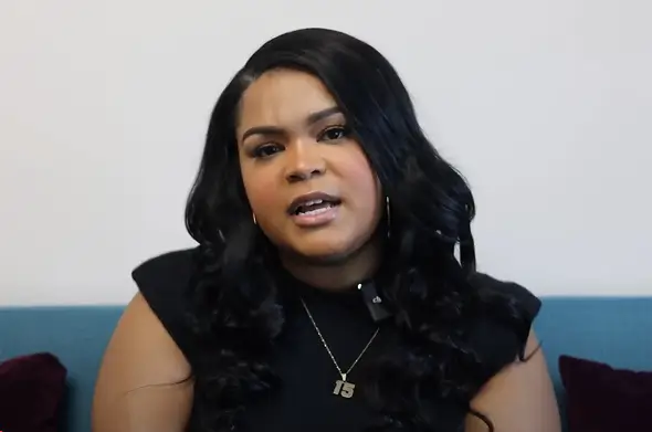 A woman with long black hair and a "15" necklace sits on a blue couch, facing the camera against a plain white background.