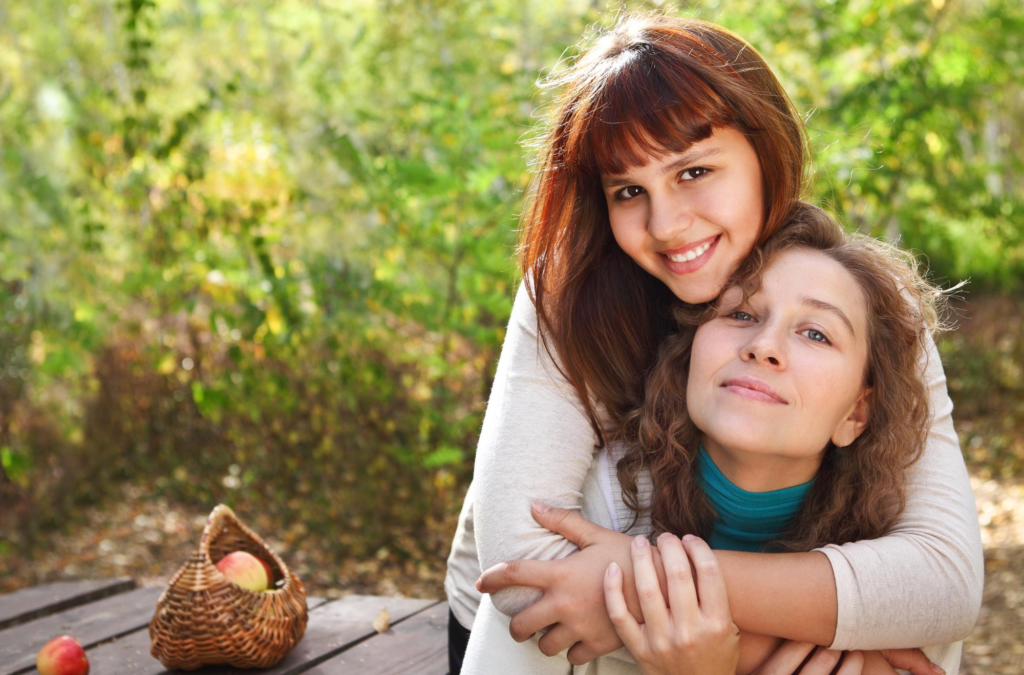 Two women sit outdoors at a wooden table. One hugs the other from behind. A wicker basket with apples is on the table. Green foliage fills the background.