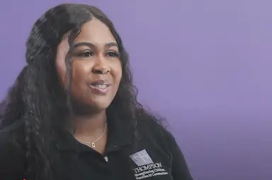 A woman with long dark hair wearing a black shirt with a "COMMISSION Community Colleges" logo sits against a plain purple background.