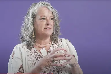 A woman with wavy gray hair and a patterned blouse gestures with her hands while speaking in front of a purple background.