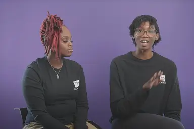 Two people sit against a purple background; one speaks while gesturing with a hand, and both wear black long-sleeve shirts with a white logo.