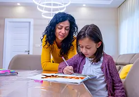 An adult woman watches a young girl draw with colored markers on paper at a table in a brightly lit room.