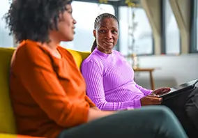 Two women sit on a couch indoors, facing each other and having a conversation. One woman holds a tablet on her lap.