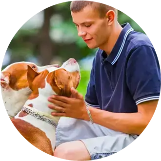 A young man sits outdoors, petting and interacting with two dogs, both looking up at him attentively.