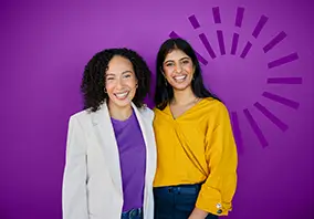 Two women stand smiling in front of a purple background with a radial graphic design. One wears a white blazer and purple shirt, the other a yellow blouse and jeans.