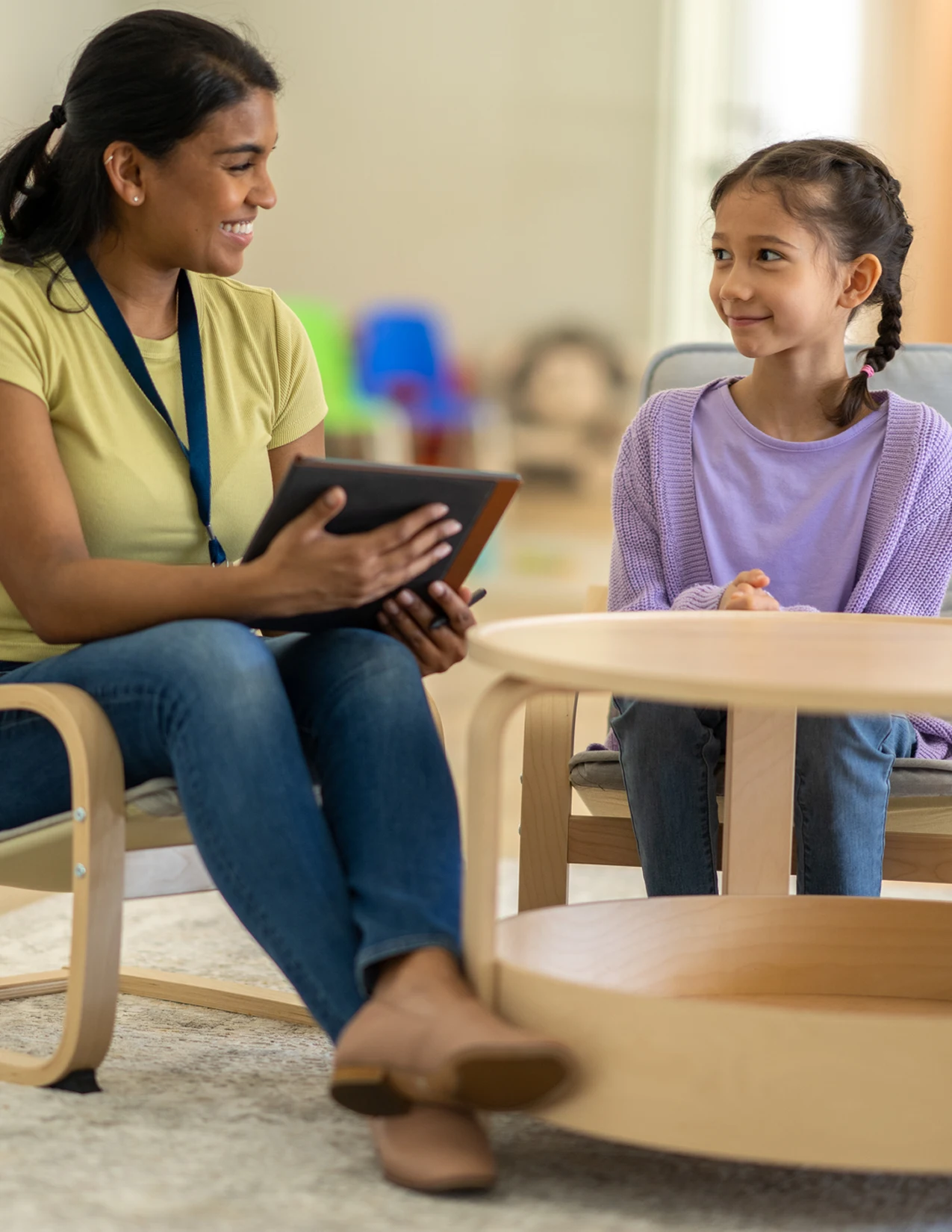 An adult woman holding a tablet talks to a young girl sitting across from her at a low round table in a bright room.