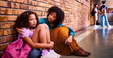 An adult comforts a sad child sitting against a brick wall in a school hallway, while other students talk in the background—reflecting Prevention efforts at Thompson Child and Family Focus.