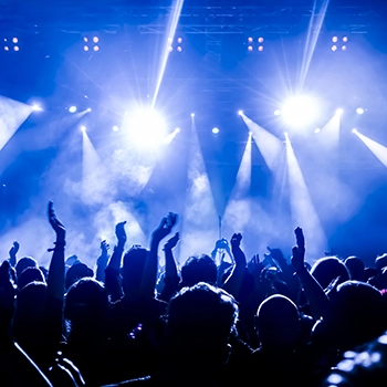 A crowd of people at a concert with raised hands, illuminated by bright blue and white stage lights and smoke effects.