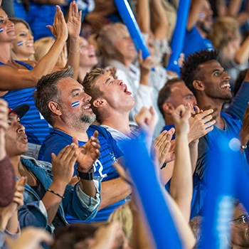 A crowd of enthusiastic fans wearing blue shirts and face paint cheer and clap at a sporting event.