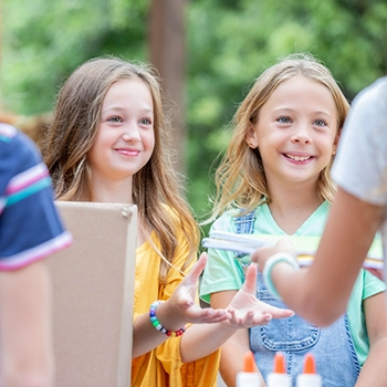 Two young girls stand at a table outdoors, smiling and reaching out while another person hands something to them. Art supplies are visible on the table.
