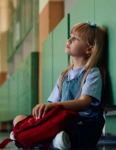 Young girl with long blonde hair sits on the floor against a green wall in a hallway, holding a red backpack and looking thoughtfully into the distance, reflecting concerns about North Carolina Medicaid rate cuts for child welfare services.