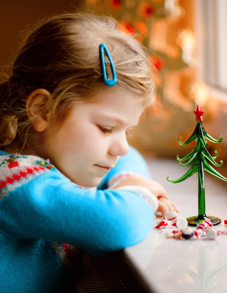 A young girl with a blue hair clip looks down at peppermint candies next to a small glass Christmas tree on a windowsill.