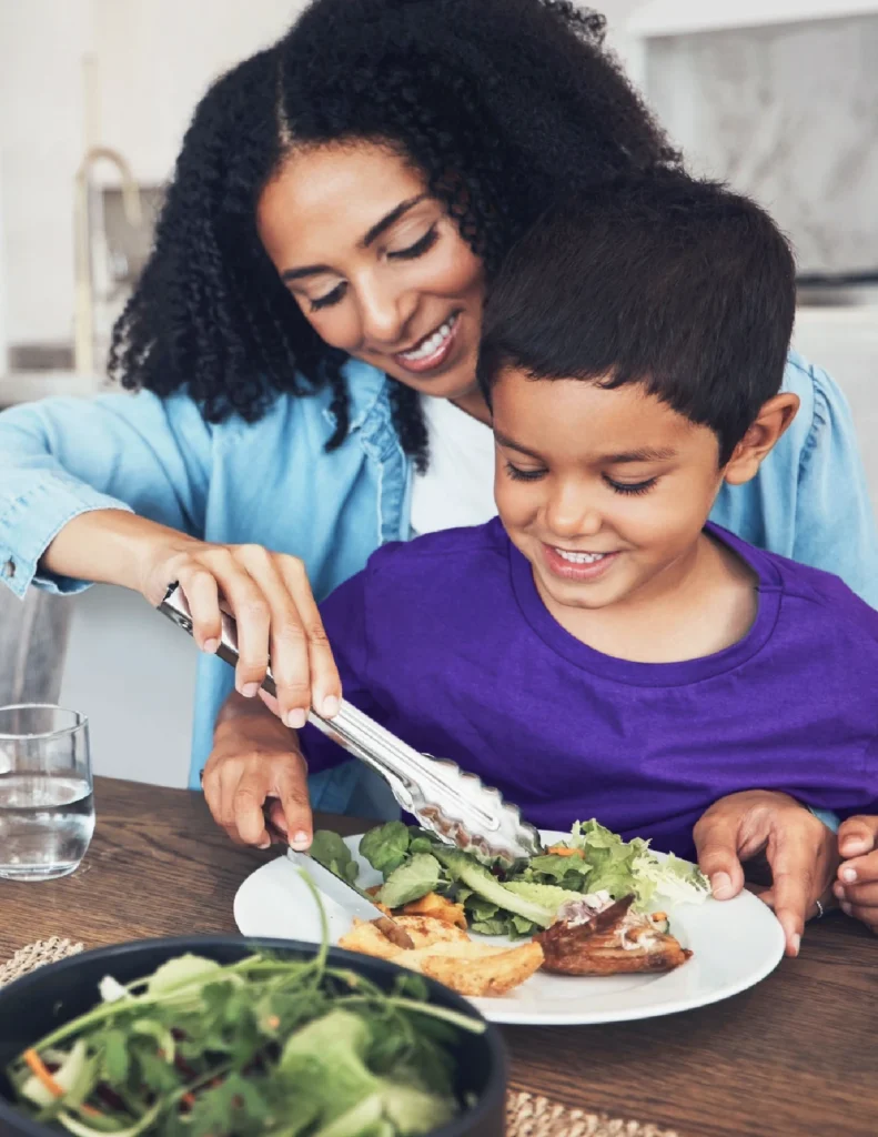 An adult and child sit at a table as the adult serves salad onto the child’s plate. Both are smiling, and there is a glass of water and a bowl of salad on the table.