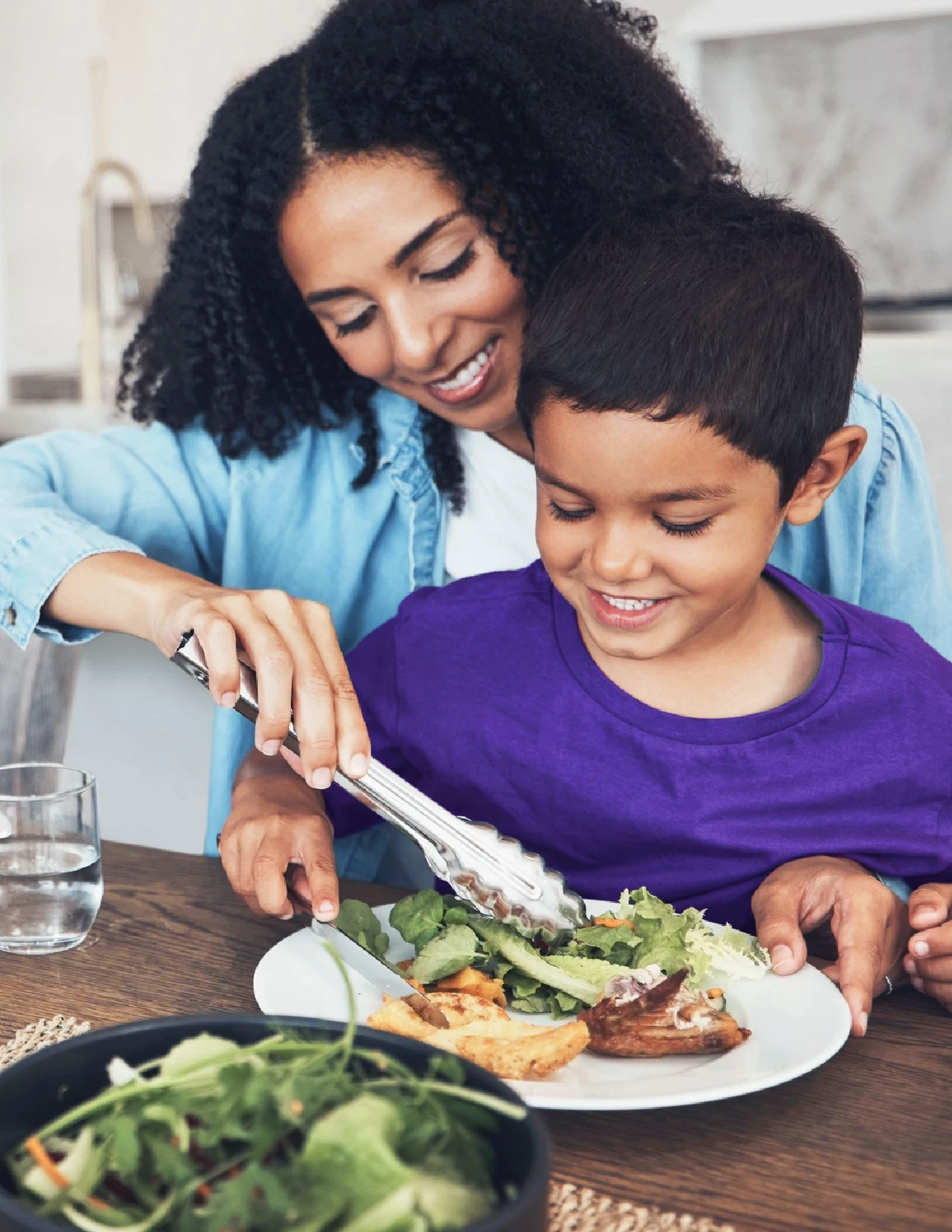 An adult and child sit at a table as the adult serves salad onto the child’s plate. Both are smiling, and there is a glass of water and a bowl of salad on the table.