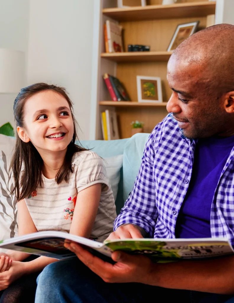 An adult and a child sit on a couch, smiling at each other while looking at an open book. A bookshelf is visible in the background.