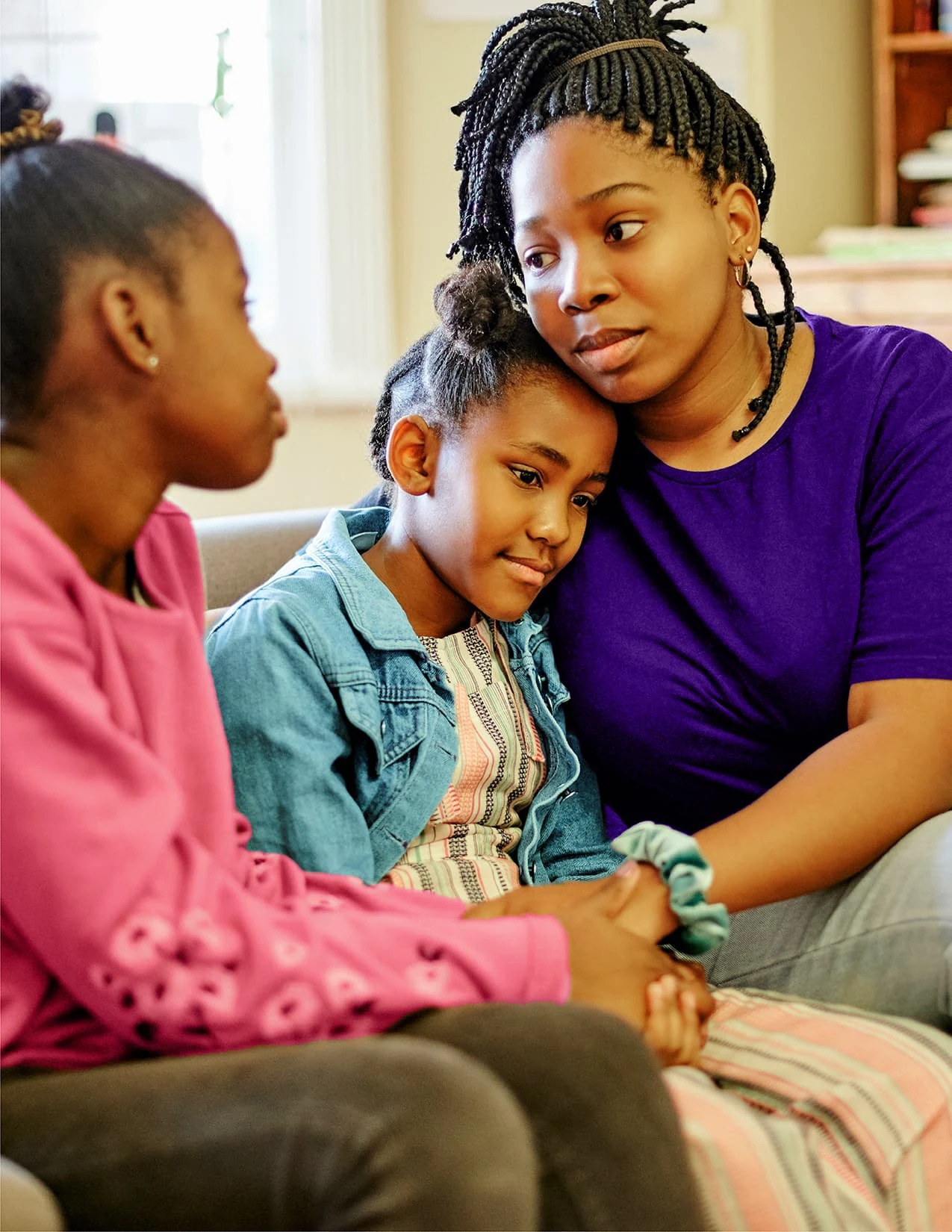 Three people sit closely together on a couch; a woman in a purple shirt hugs a young girl while another girl sits nearby, all appearing to have a serious conversation, strengthening families through support and understanding.