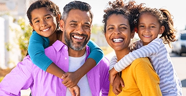 Smiling family of four standing outdoors, with two children hugging their parents from behind, embracing prevention and support from Thompson Child and Family Focus.