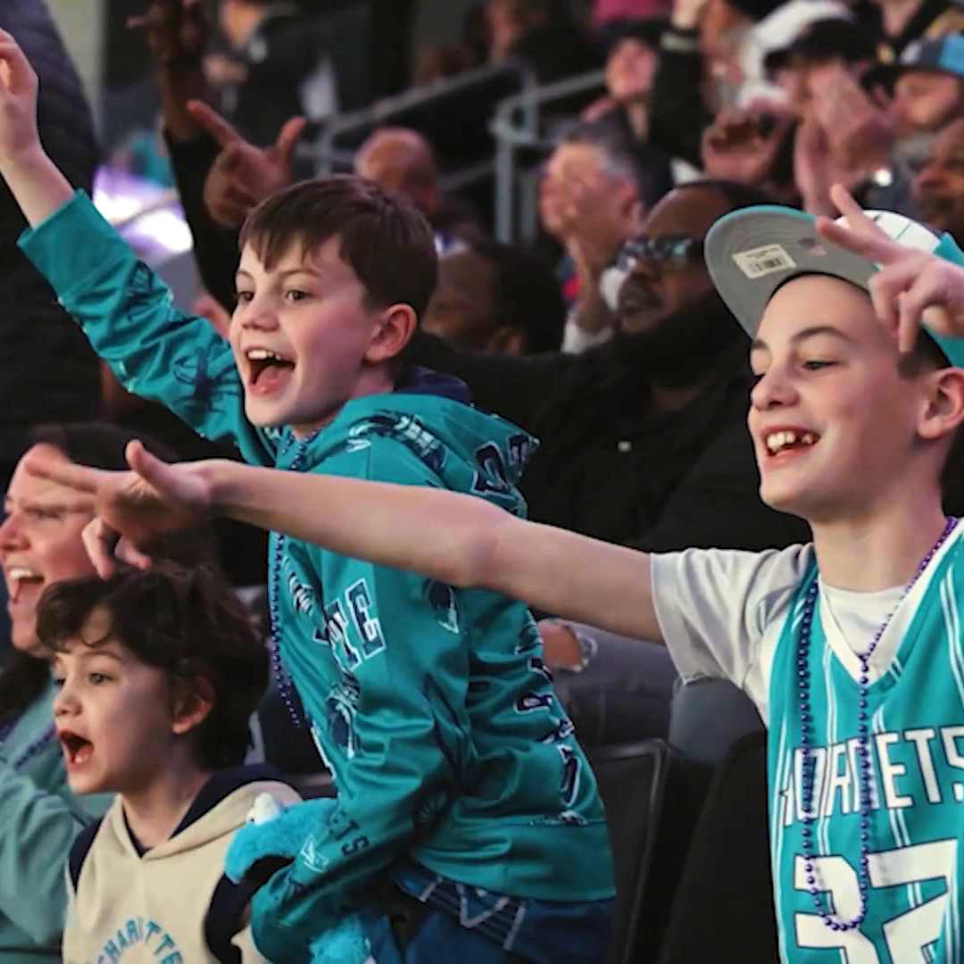 Three excited children wearing teal Charlotte Hornets gear cheer and gesture enthusiastically at a basketball game, with a crowd visible in the background.