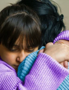 A young girl with bangs hugs an adult wearing a blue sweater, both dressed in knit clothing, showing a close and comforting embrace.