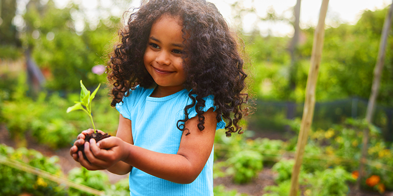A young girl with curly hair holds a small plant in her hands while standing in a garden, symbolizing how even a small donation can help nurture growth and positive change.