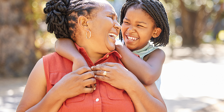 An older woman and a young girl smile and embrace outdoors, with the girl’s arms around the woman’s shoulders. Their happiness shines as they share a special moment, reminding us how every donation can help bring joy to families like theirs.