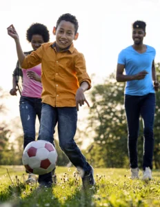 A boy in an orange shirt kicks a soccer ball on grass while two adults run behind him on a sunny day.