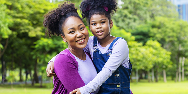 An adult and a child smiling and posing together in a park with green trees in the background, celebrating the joy that comes from giving through donation.