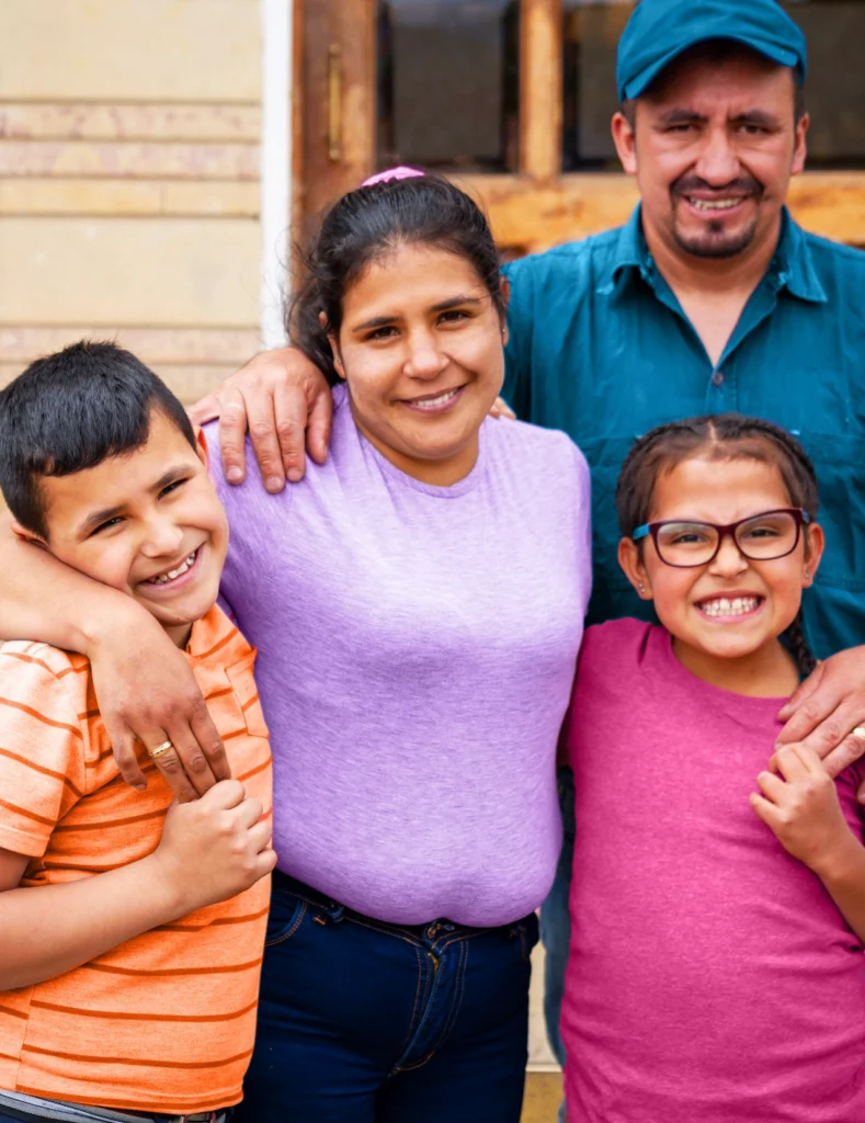 Two adults and two children stand close together outdoors, smiling at the camera. The woman and children wear colorful shirts; the man wears a blue shirt and cap.