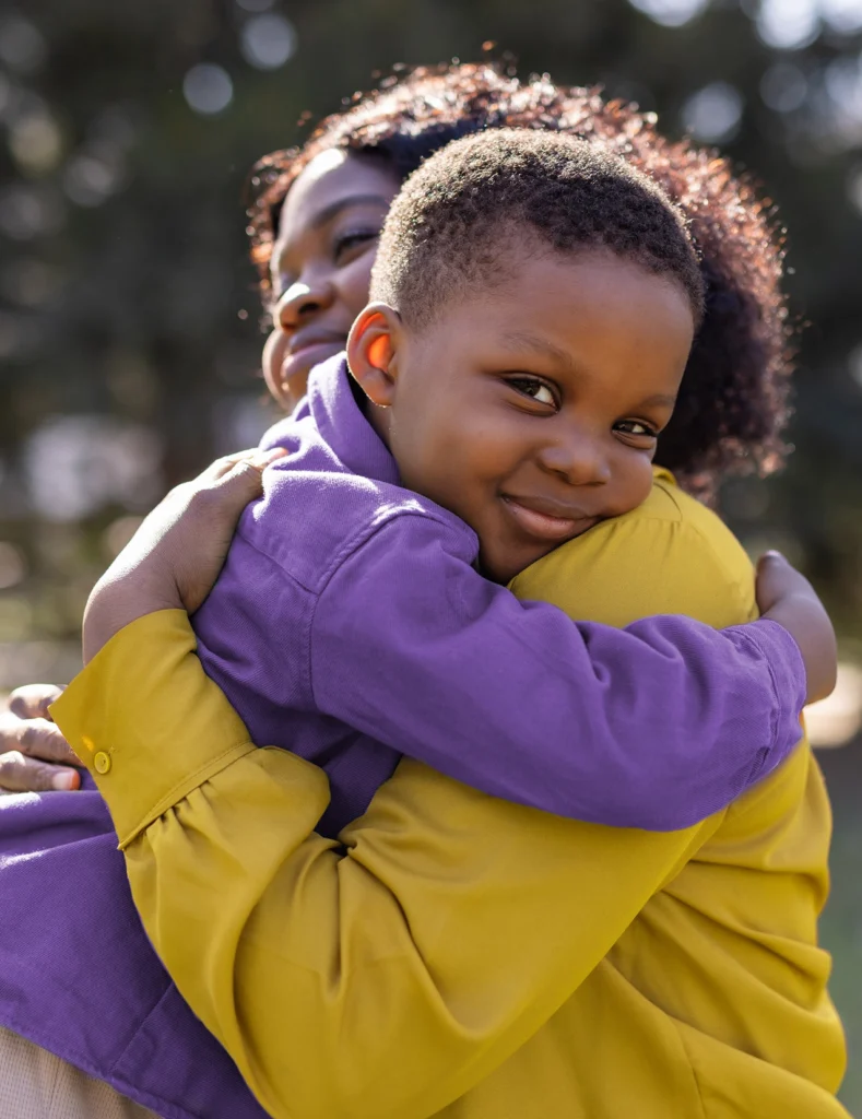 A young child in a purple shirt hugs an adult in a yellow jacket outdoors, both smiling warmly.