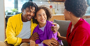 Three people sit together on a couch in a living room, smiling and talking. The person on the left wears yellow, the middle person wears purple, and the right person wears red. Their warmth and connection reflect the supportive environment foster care can create—a core commitment of Thompson Child & Family Focus.