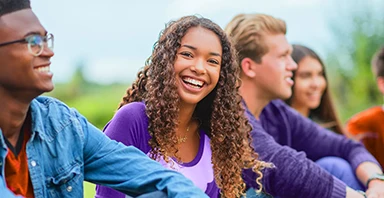 A group of young adults sit outdoors on grass, smiling and talking together, representing the supportive community fostered by Thompson Child & Family Focus. The focus is on a woman with curly hair wearing a purple shirt, highlighting the positive connections formed through foster care.