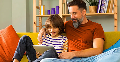 A man and a child sit on a yellow couch looking at a tablet together, smiling—sharing a moment of connection that reflects the supportive environment fostered by Thompson Child and Family Focus’s mental health services. Books and decorative items are on a shelf in the background.