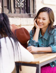 Two women sit at an outdoor table talking. One woman, smiling, faces the camera, while the other’s back is turned. A brown hat rests on the table beside them.