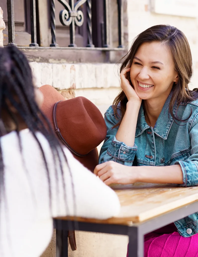 Two women sit at an outdoor table talking. One woman, smiling, faces the camera, while the other’s back is turned. A brown hat rests on the table beside them.